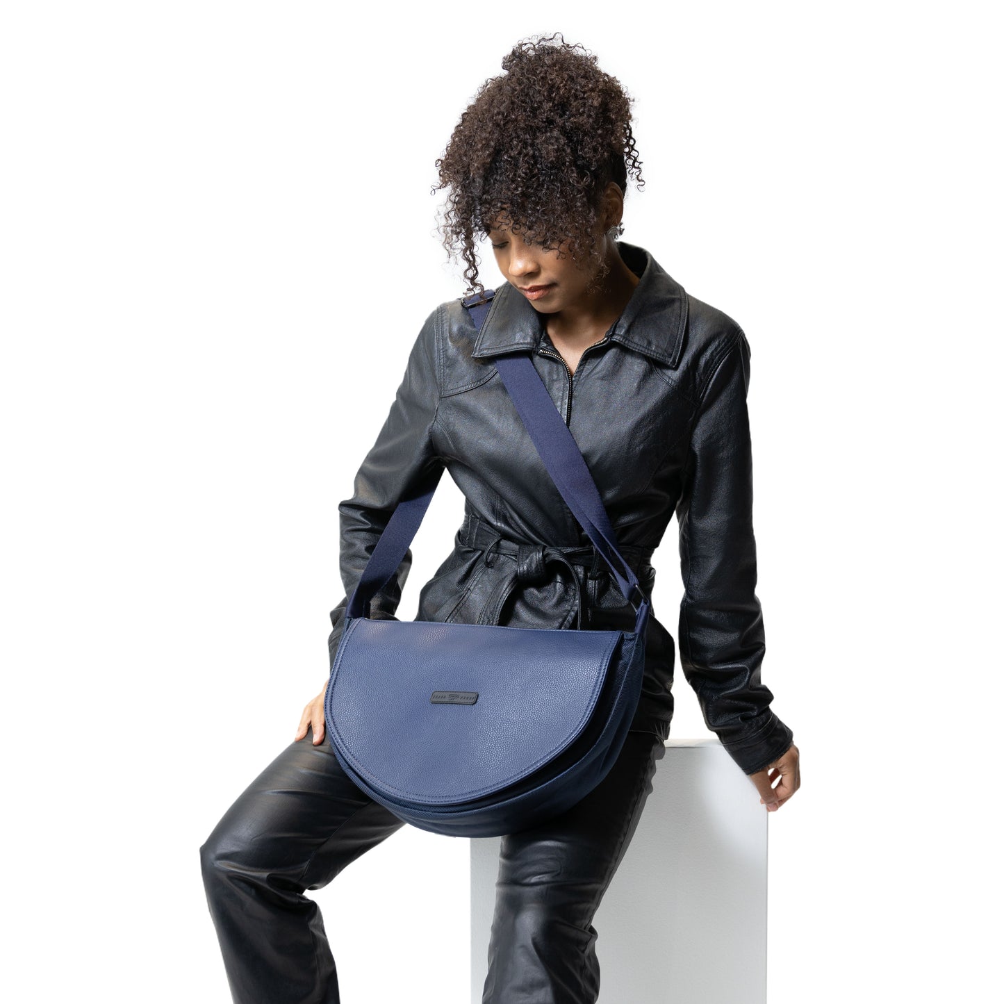 Model wearing a black leather outfit sitting in a white background wearing a blue smell proof crossbody bag