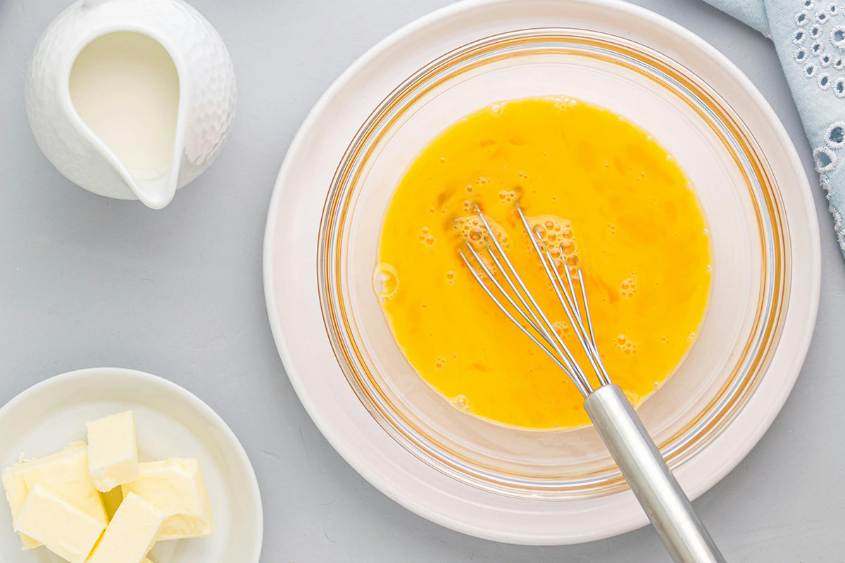 Eggs being whisked in a bowl with a whisk and butter on the side on a light gray surface.