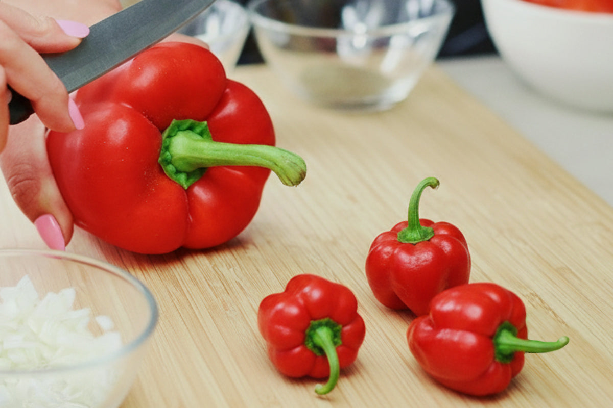 Red bell pepper being cut on a wooden cutting board with small red peppers and chopped onions.