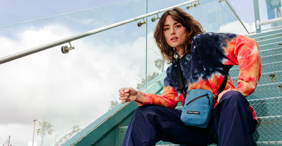 Woman sitting on steps wearing a tie-dye jacket and holding a blue Stash Bag.