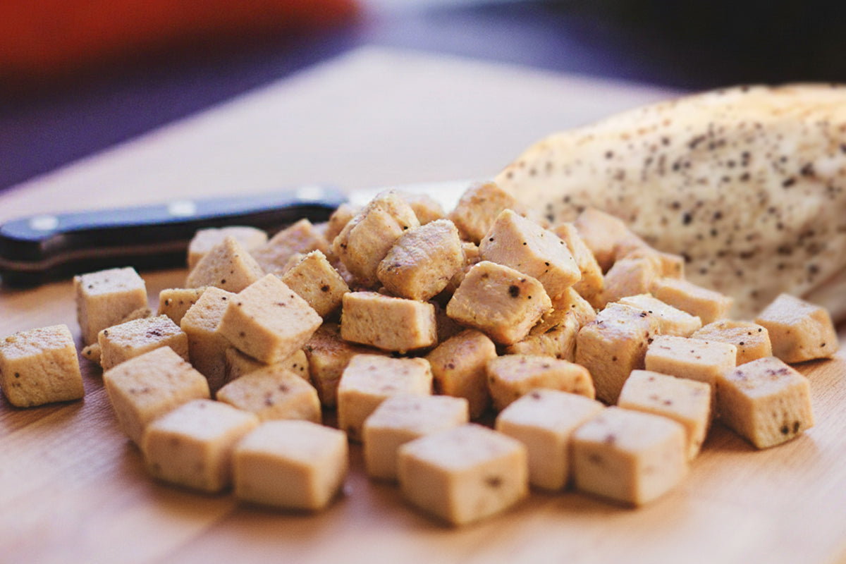 Cubed chicken on a wooden cutting board with a knife and bread in the background.