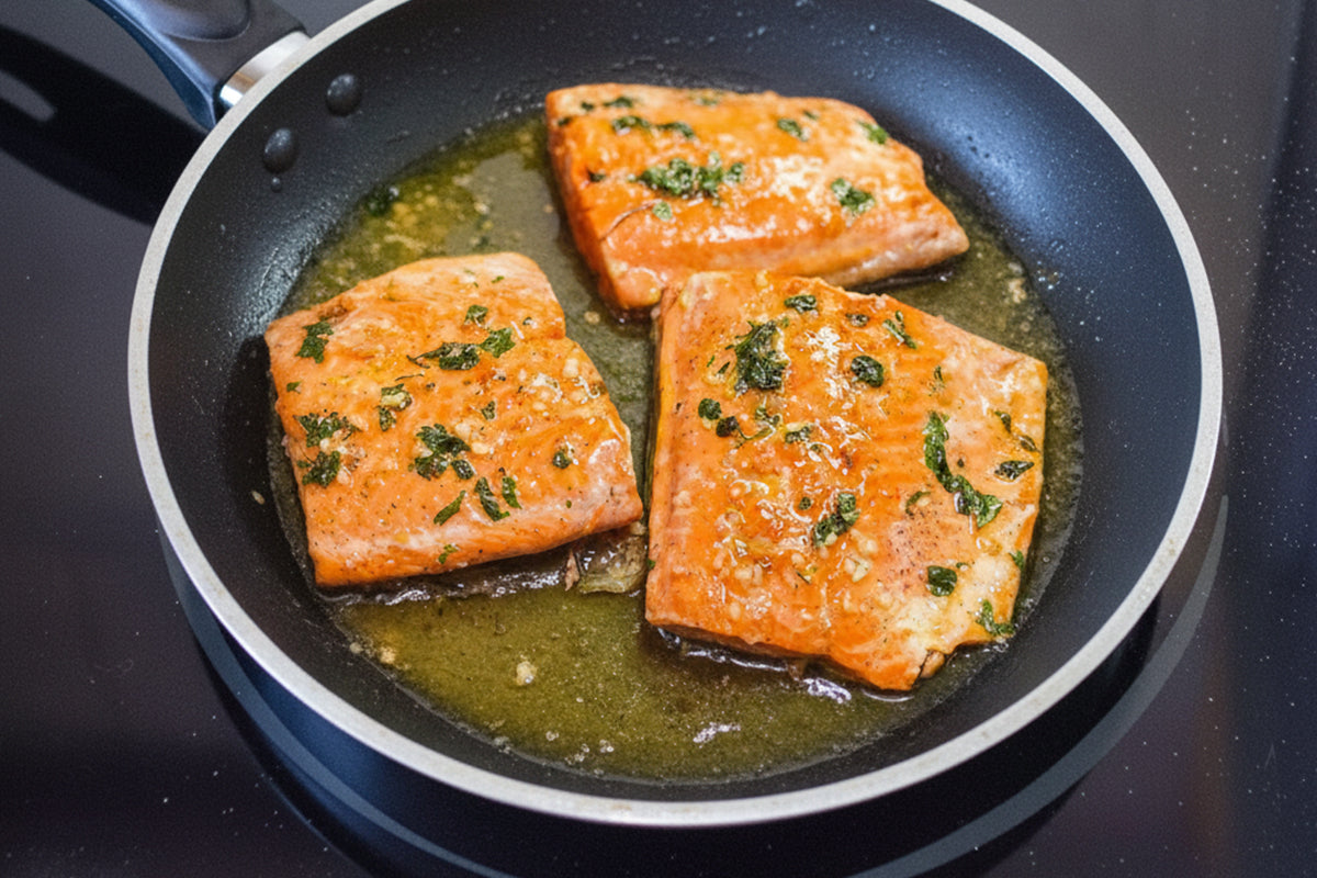 Three pieces of salmon cooking in a pan on a stovetop.